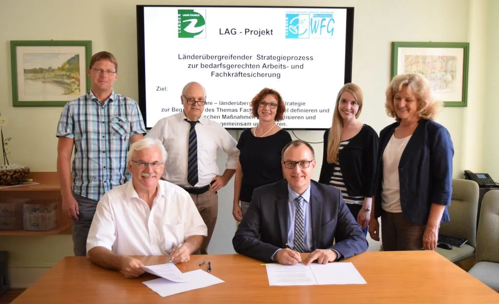Der Erste Kreisbeigeordnete Helmut Jung (links) und Bürgermeister Michael Schnatz (rechts) unterzeichnen den Kooperationsvertrag. Im Hintergrund stehend von links nach rechts die beteiligten LEADER-Regionalmanagements: Lars Wittmaack, Walter Gerharz und Martina Donnert für die Region Limburg-Weilburg sowie Carolin Stoeppel und Sabine Ksoll für die Region Lahn-Taunus. Foto: Leader Lahn Taunus